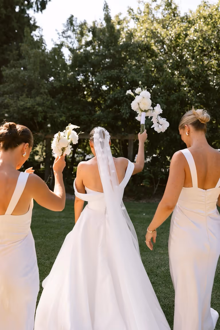 A bride in a white dress and her bridesmaids walk across the lawn holding white bouquets
