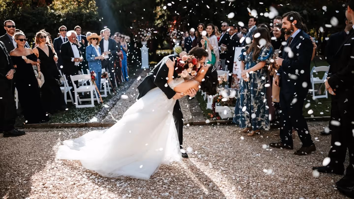 A bride and groom share a kiss as guests throw petals on the gravel path at Fitzroy Inn